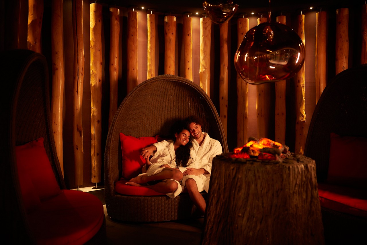 Man and woman snuggled up in a large cocoon chair by a glowing fire.